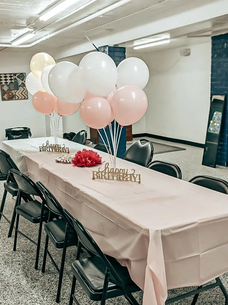 Long table with folding chairs decorated for a birthday party with light pink and white tablecloths, pink and white balloons, and small standees that say "Happy Birthday!" in gold script text.