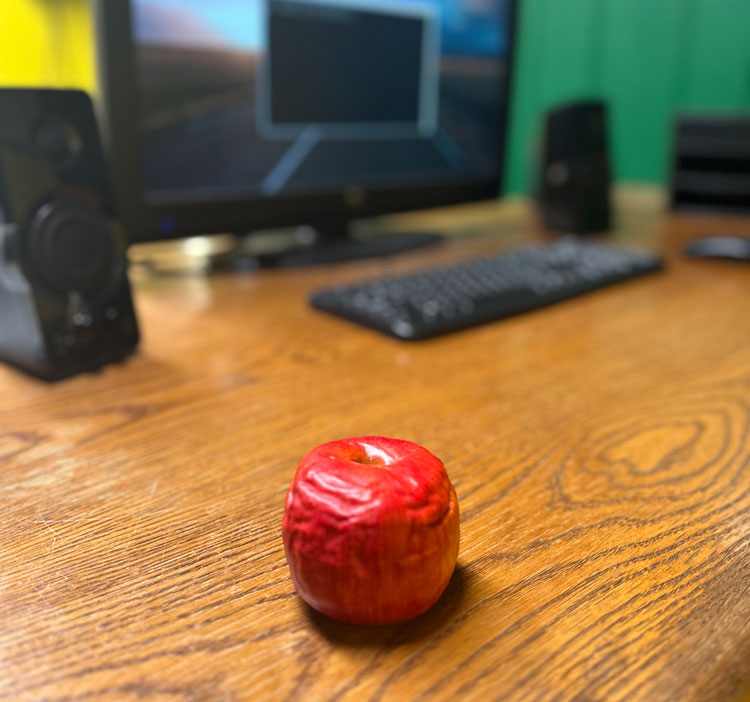 A red apple sitting on a desk with an out of focus computer in the background.