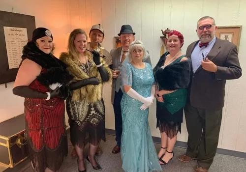 A group of 7 people dressed up in 1920s era clothes for a murder mystery party.